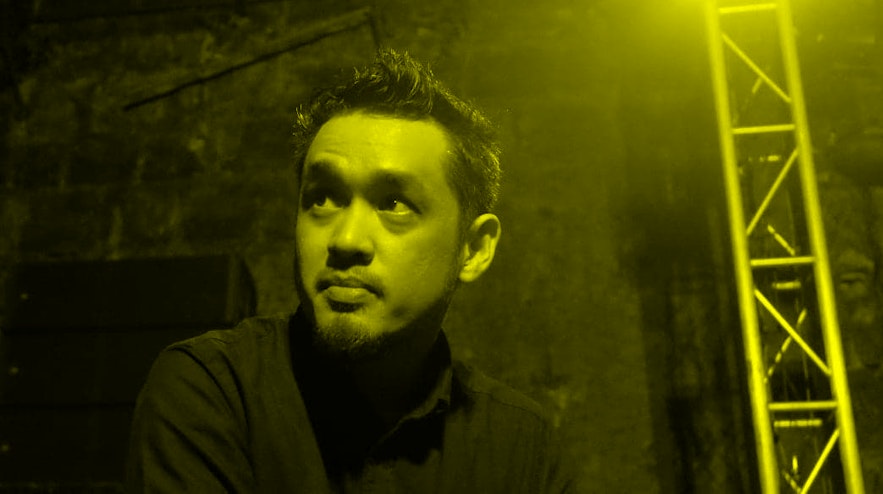 A person with short black hair and light brown skin raises their eyebrows an looks up. They are wearing a black turtleneck and sitting in front of a stage.