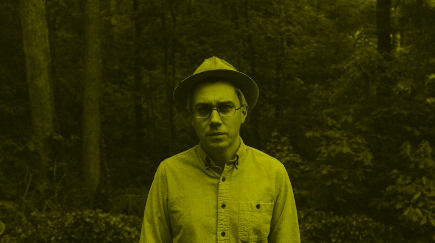 A person with light skin and short grey hair wearing a brimmed hat stands in shady a forest glade. They have a slightly concerned expression.