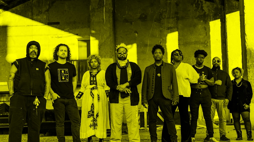 Nine people standing in a row with serious expressions. Most of them have short, curly black hair and their skin tones range from light to dark brown.