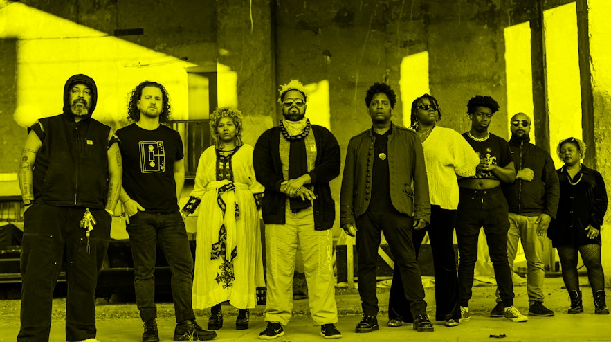 Nine people standing in a row with serious expressions. Most of them have short, curly black hair and their skin tones range from light to dark brown.