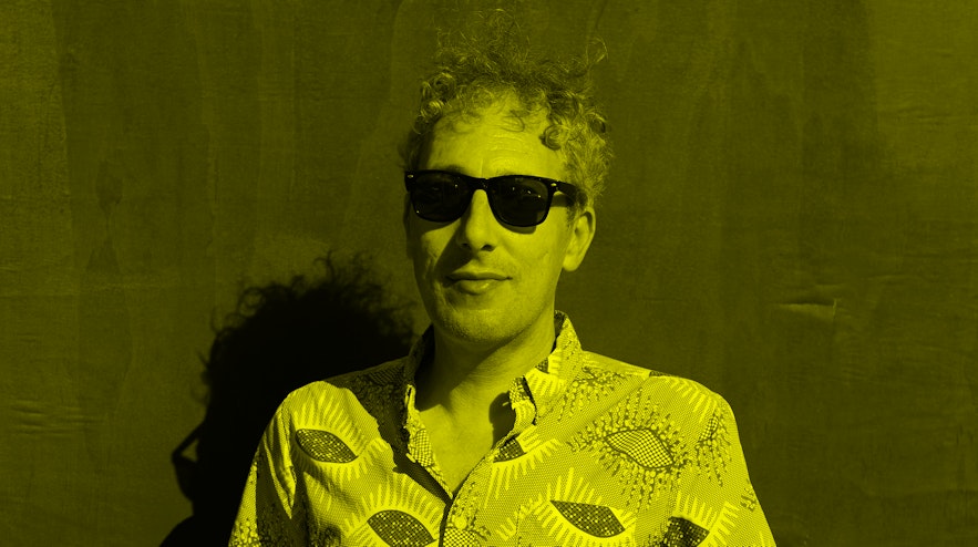A person with short, grey, curly hair and light skin wearing sunglasses. They have a hint of a smile.