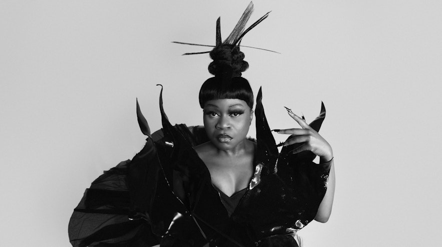 A person with straight black hair and a fringe and medium brown skin wearing an elaborate black gown is viewed from above, looking directly at you.