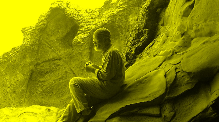 A person with a white beard and light skin sits on a sandstone formation with their hands clasped. They are viewed from the side.
