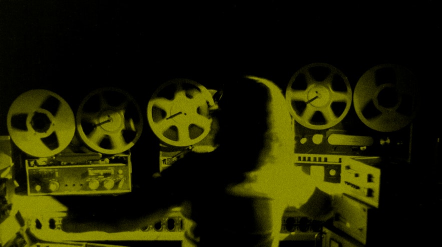 A person with light, long hair is viewed from behind. Their arms are stretched across several mixing desks. Above them are six large tape reels.