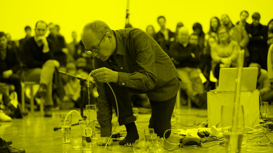 A person with short grey hair and light skin and glasses bends over several glasses of water on the floor. They are dangling a tube with a microphone into one of the glasses.