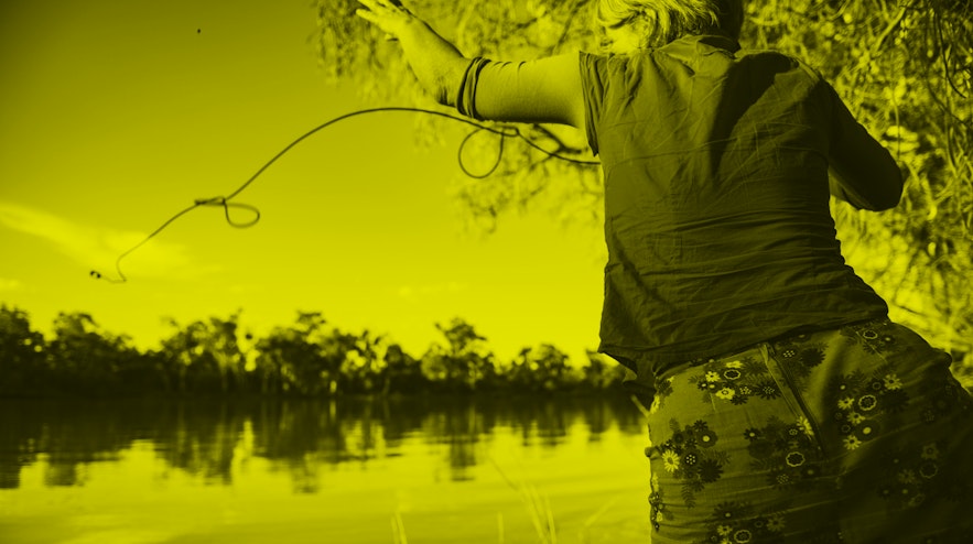A person with short grey, brown hair and light skin flings a microphone on a rope into a river on a sunny day.