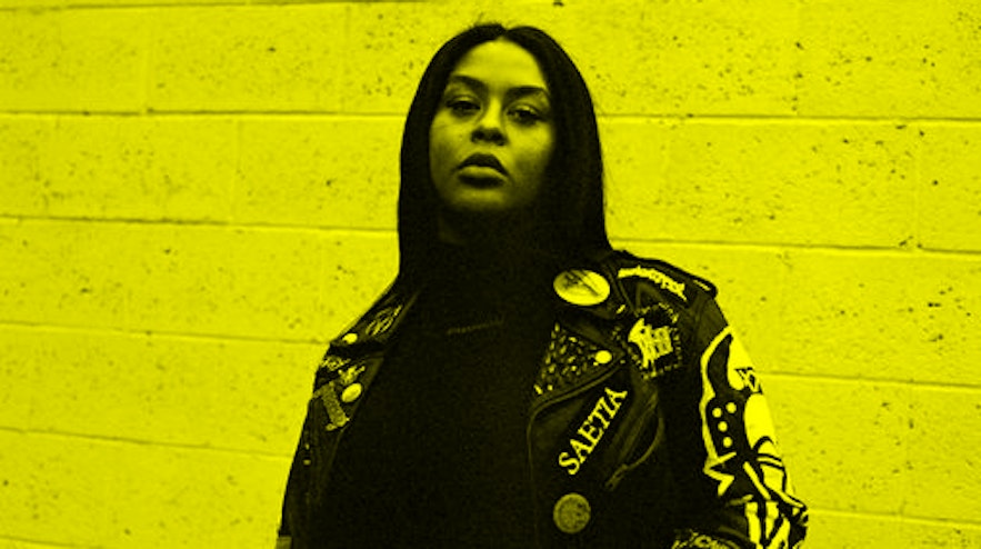 A person with long black hair and medium brown skin stands with one hand on their jacket.