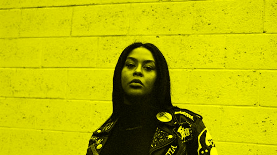 A person with long black hair and medium brown skin stands with one hand on their jacket.
