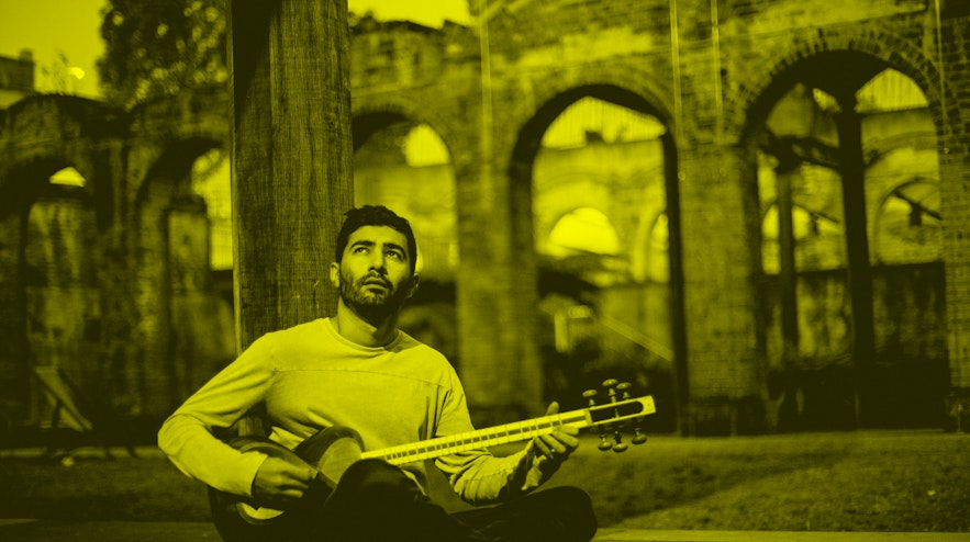 A person with short black hair and light brown skin seated on the ground playing a tar.