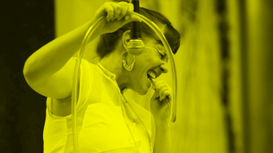 Person in white coveralls, holding a long transparent tube in one hand and holding the end of it to their mouth in the other.