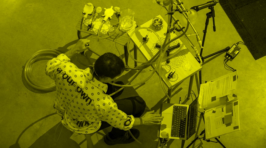 Person, framed from above, sitting on a stool, surrounded by three small tables with various tubes and wires.