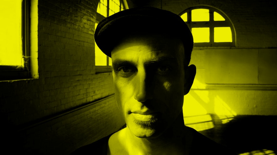 Person pictured from the neck upwards, in a dark room, wearing a flat cap and staring at the camera openly.