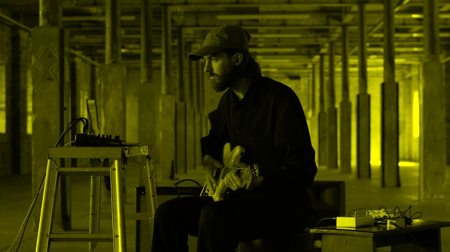 Person sitting and playing an electric guitar, with various digital equipment on tables either side.