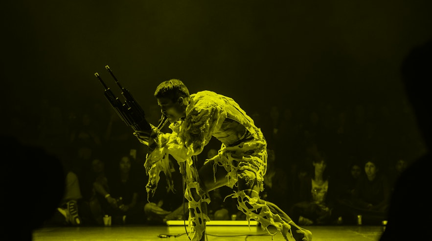 Person crouching wearing a ragged white outfit, blowing into a two pronged electrical instrument