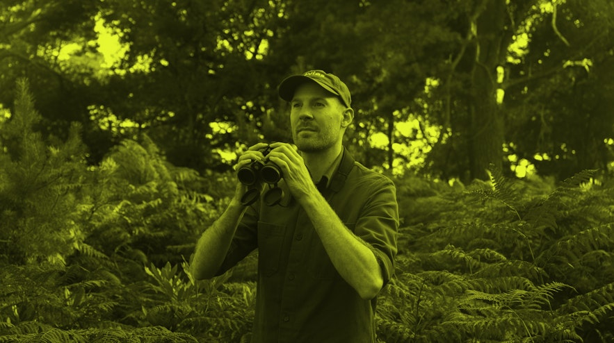 A person with light skin wearing a baseball cap holds a pair of binoculars. Behind them are ferns and treees.
