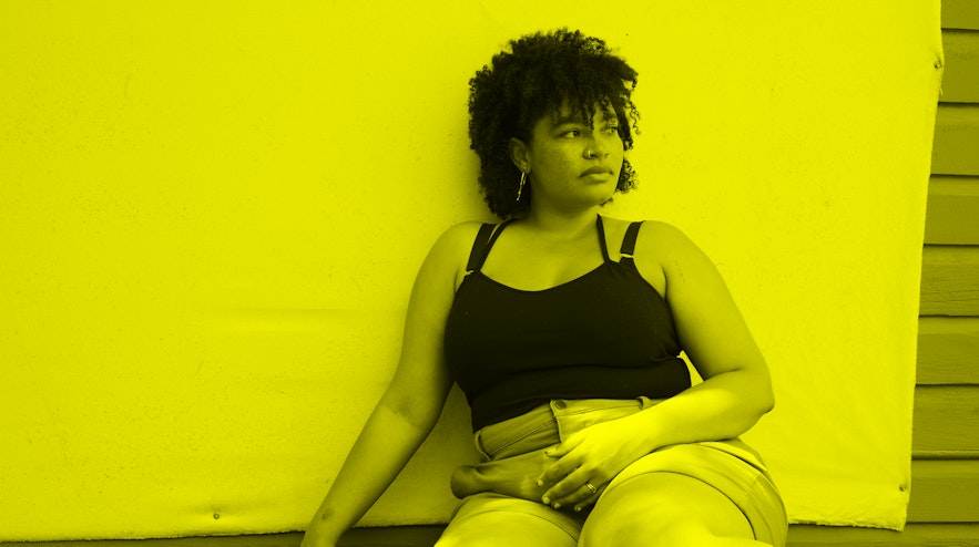 A person with light brown skin and curly black ringlets sits leaning up against a piece of canvas tacked onto a wall.