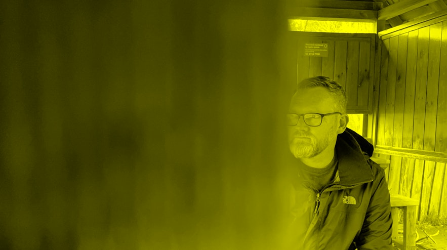 Person, image partially obscured by wooden doorway, sitting in a wooden hut